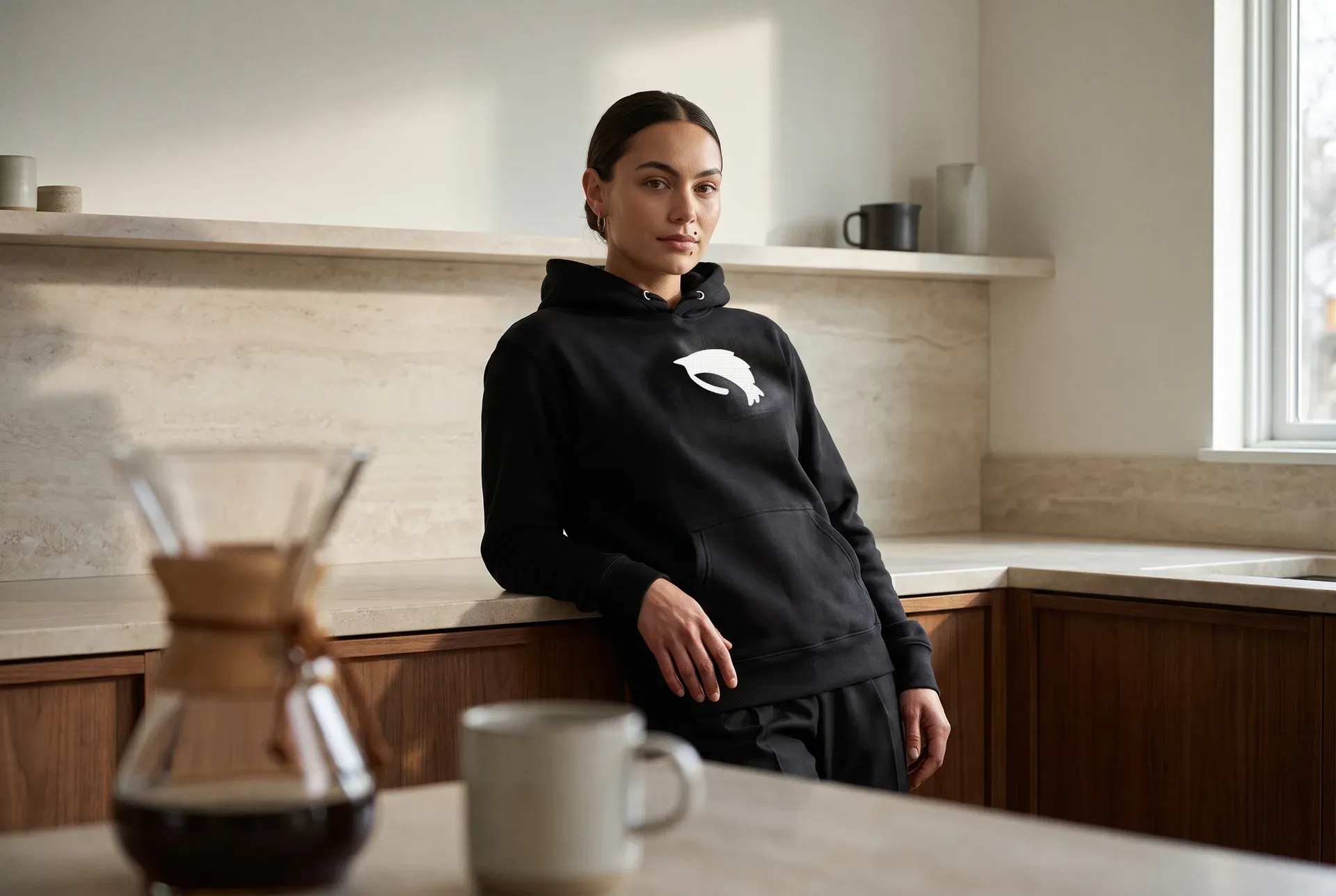 Eine Frau in einem dunklen Hoodie mit einem gestickten Garnelen-Motiv, in einer hellen Küche — ruhige, getragene Aufnahme.