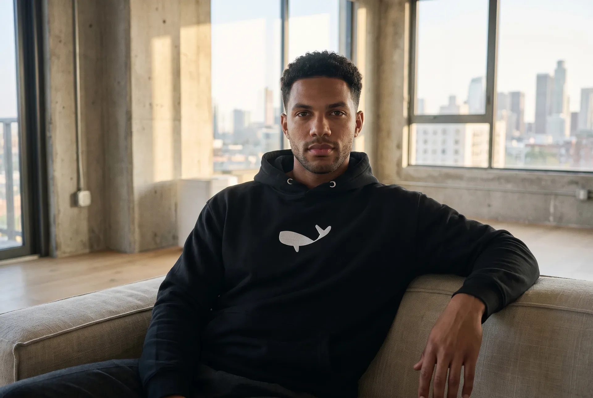 Ein Mann in einem dunklen Hoodie mit einem gestickten Buckelwal auf der Brust, in einem hellen Loft — ruhige, getragene Aufnahme.