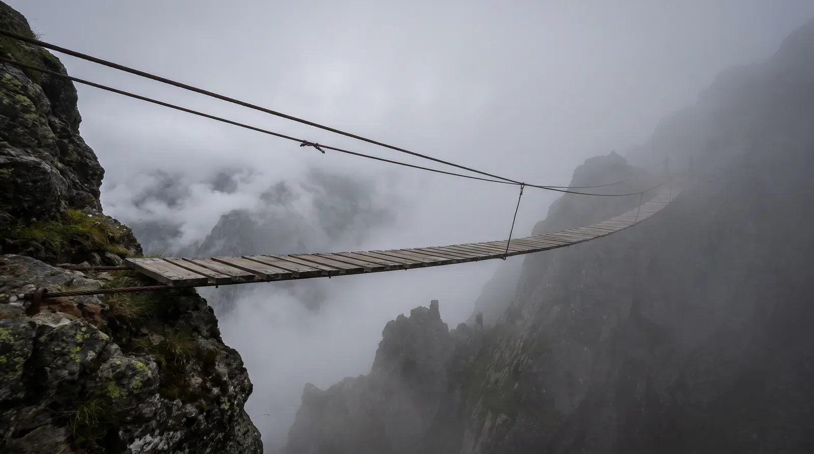 Eine schmale Hängebrücke aus Holzplanken führt über eine nebelverhangene Schlucht, das andere Ende verschwindet im Grau