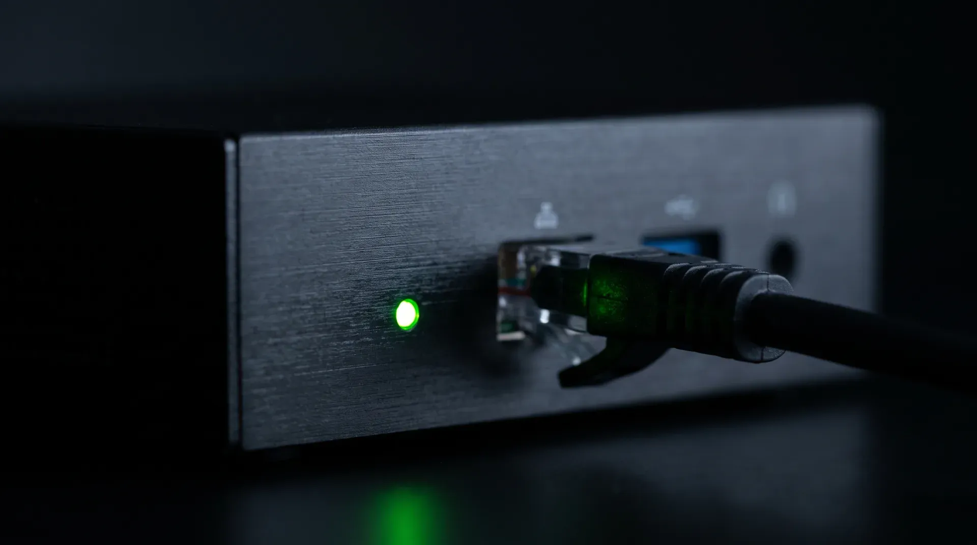 Makroaufnahme eines kleinen schwarzen Mini-Computers im Dunkeln: eine einzelne grün leuchtende Status-LED, daneben ein eingestecktes Netzwerkkabel